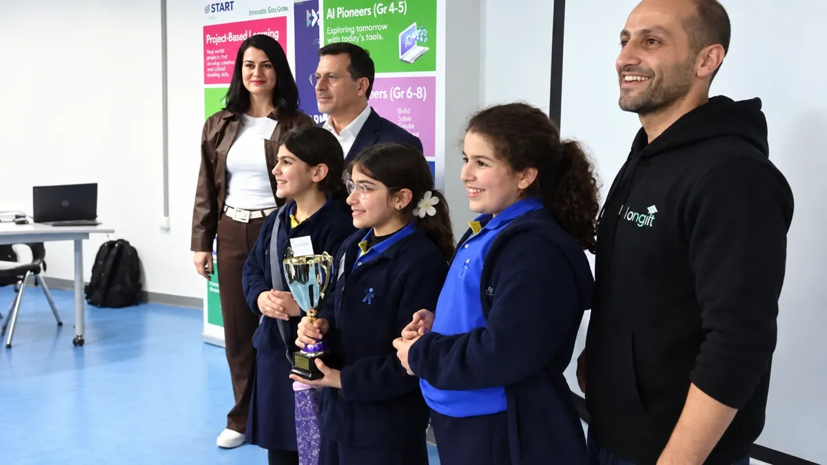 Three students in school uniform holding a trophy, standing with organisers in front of aiSTART banners at the hackathon